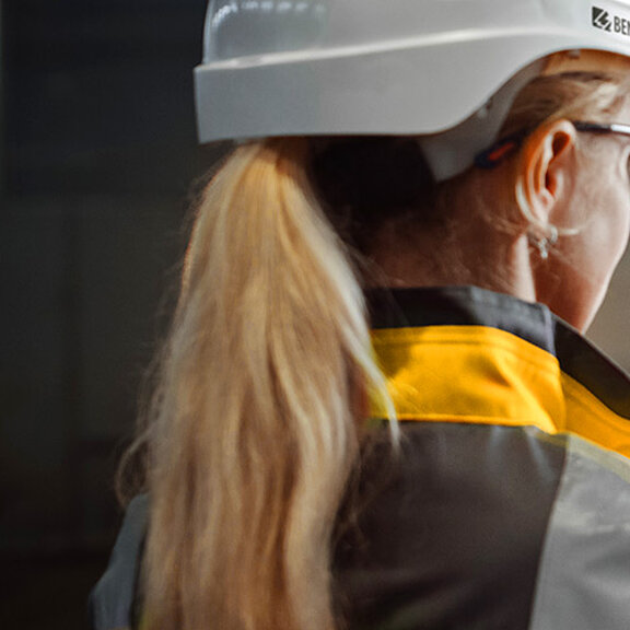 Person mit weißem Schutzhelm und gelber Arbeitsjacke hält Tablet mit technischen Diagrammen in einer Industriehalle